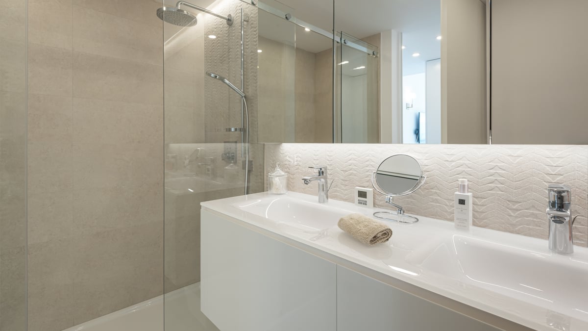 Salle de bain contemporaine avec double vasque blanche, douche vitrée et carrelage beige texturé.