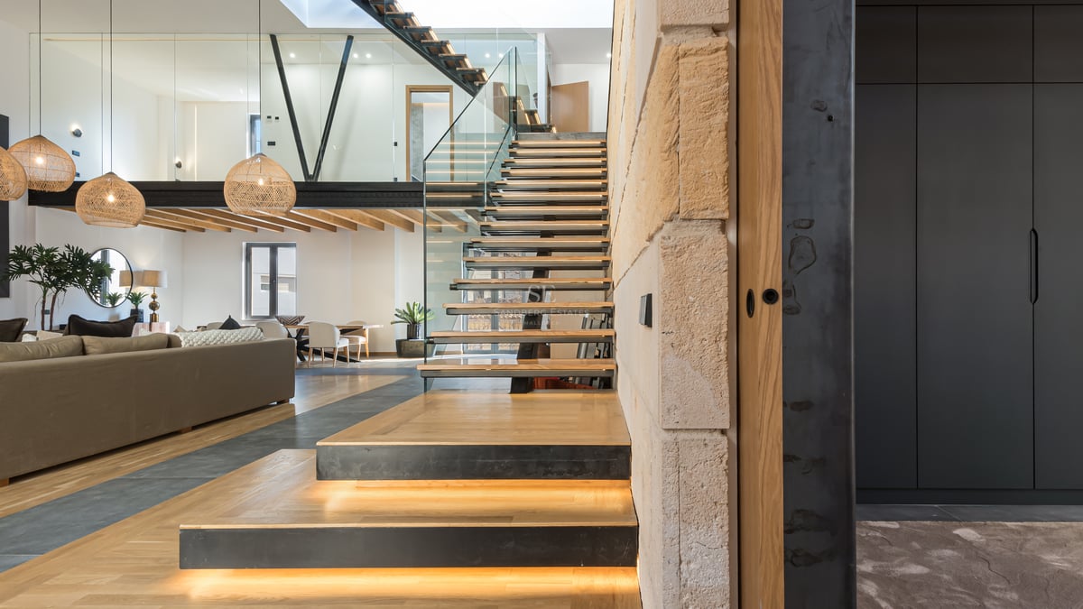 Salon moderne ouvert avec escalier flottant en bois, éclairage LED, garde-corps en verre et mezzanine.