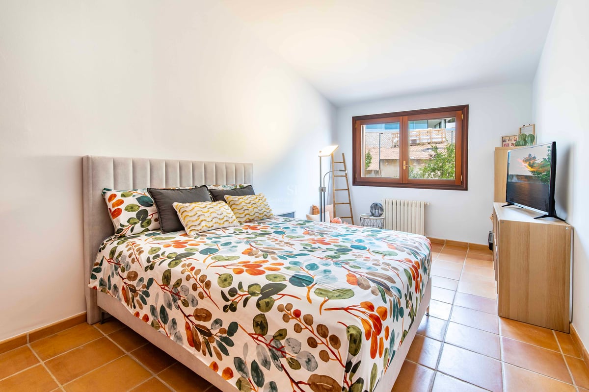 Chambre avec lit à motifs floraux, tête de lit grise, sol en terre cuite et fenêtre en bois.