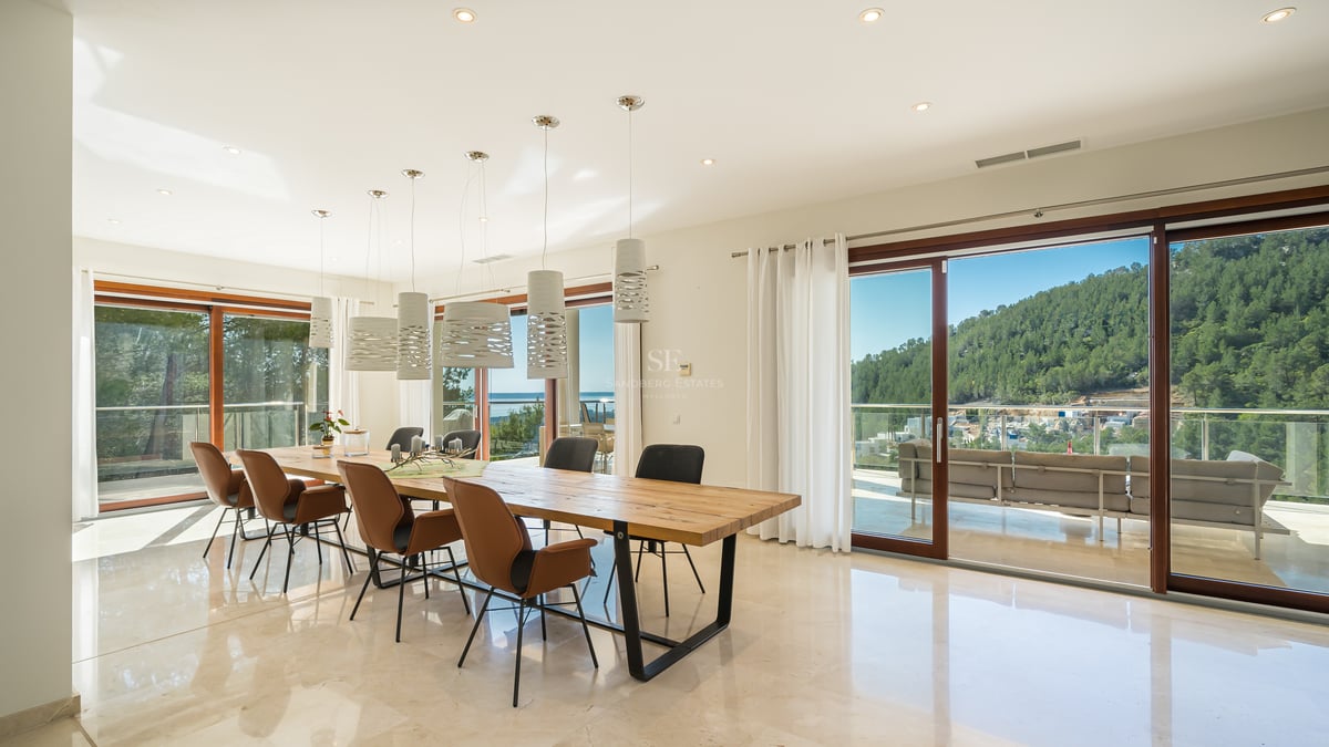 Salle à manger moderne avec table en bois, sol en marbre et baies vitrées ouvrant sur une vue montagne et mer.
