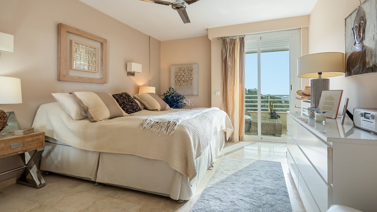 Chambre lumineuse avec lit crème, sol en marbre et grande baie vitrée ouvrant sur une terrasse ensoleillée.