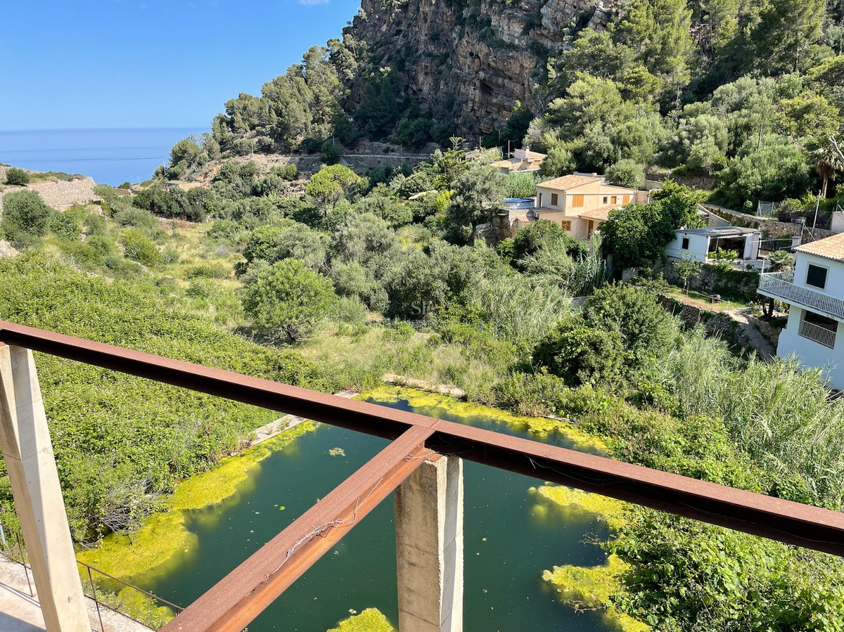 Vue depuis une terrasse en acier surplombant une vallée verdoyante avec des maisons méditerranéennes et des falaises.