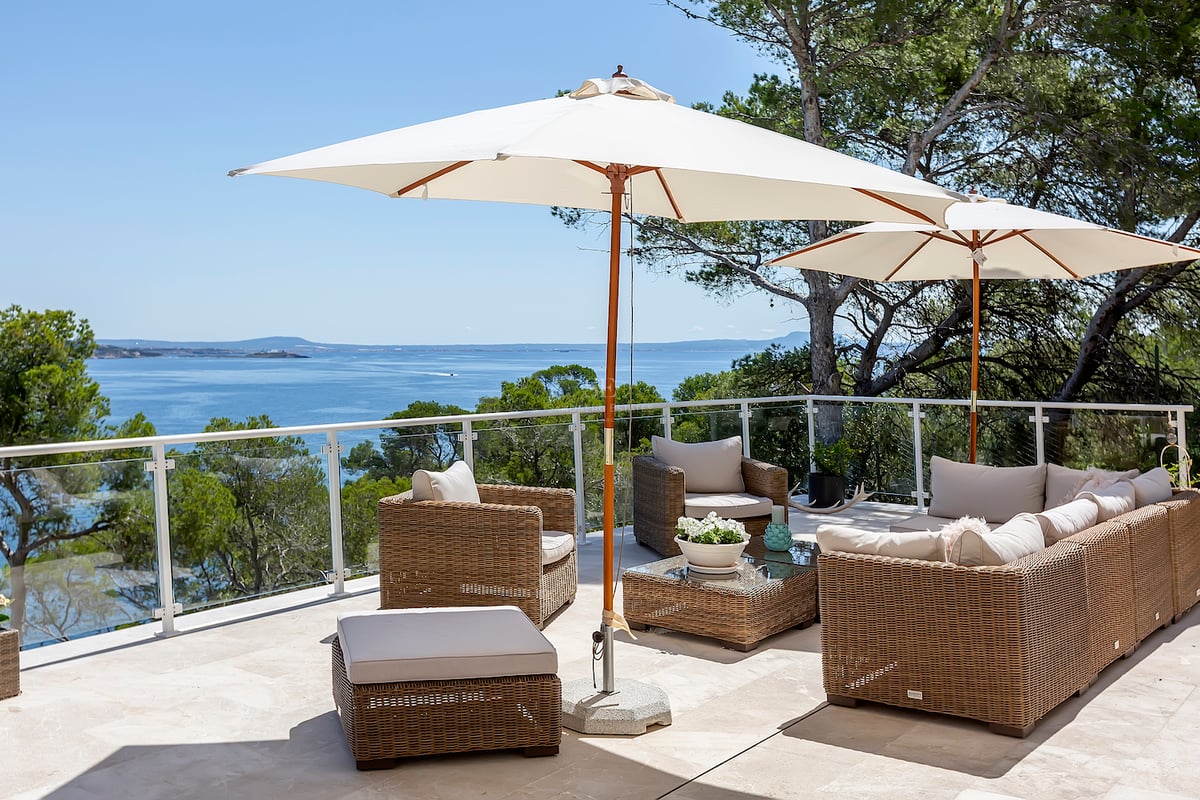 Terrasse ensoleillée avec salon en rotin, parasols blancs et garde-corps en verre surplombant la mer bleue.