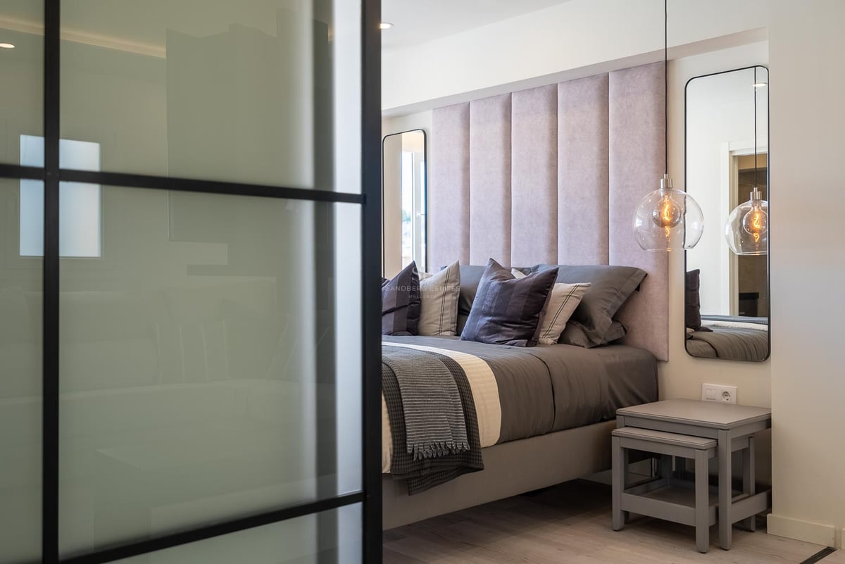 Gros plan d'une chambre principale avec tête de lit capitonnée rose, suspensions en verre et cloison industrielle.