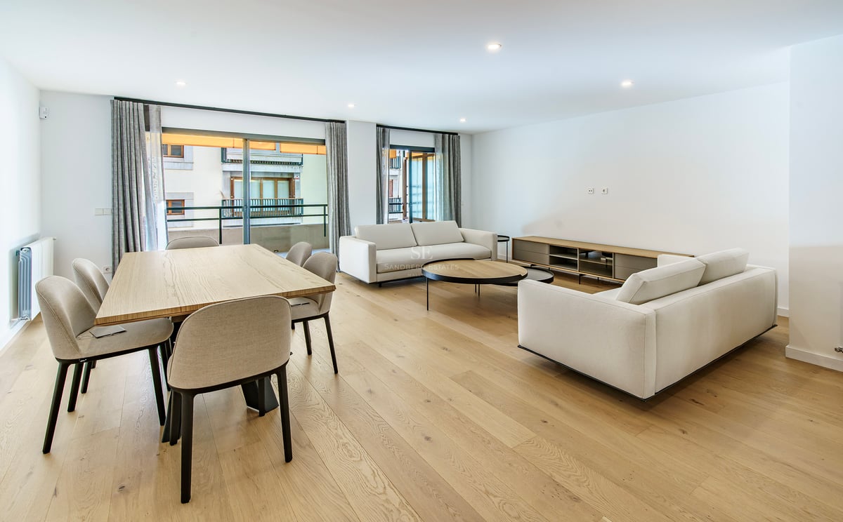 Salón abierto con sofás blancos, mesa de comedor de madera y grandes ventanales a un balcón.