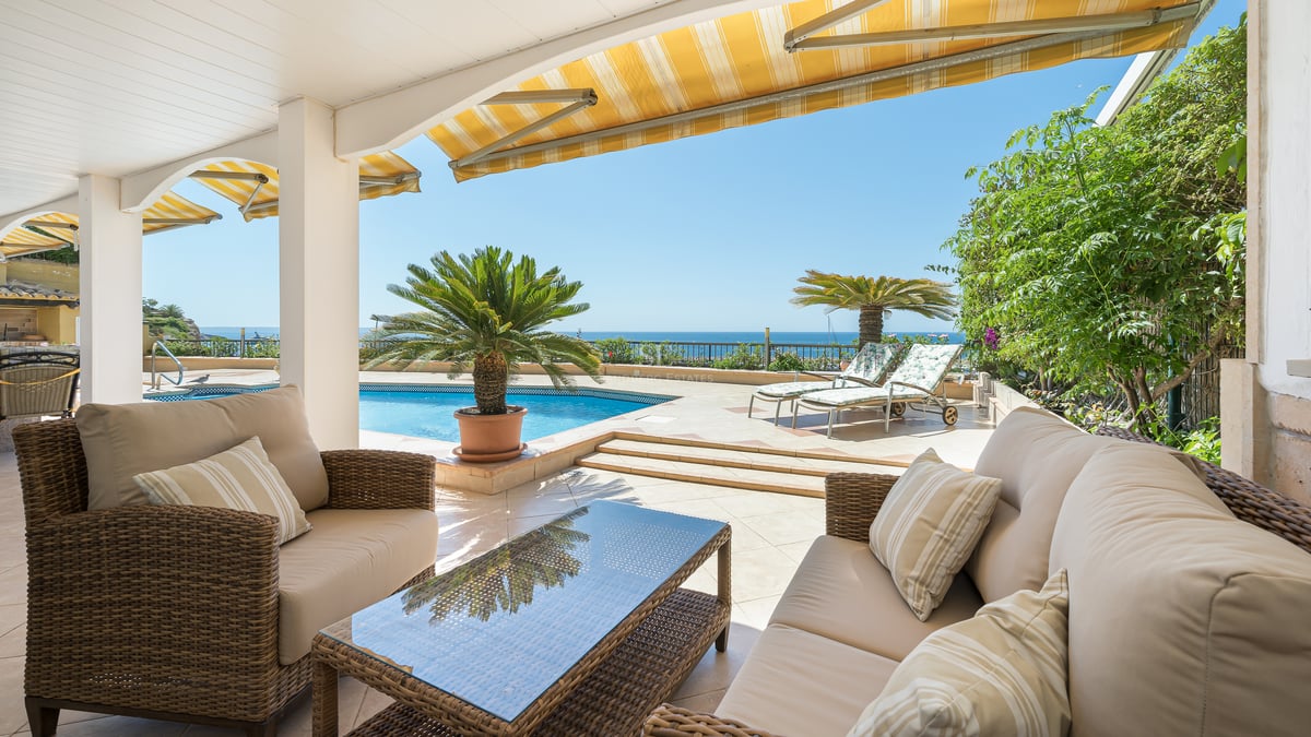Terrasse couverte avec canapés en osier face à une piscine bleue et la mer sous un ciel dégagé.