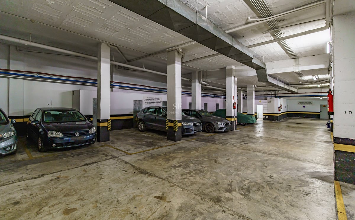 Amplio garaje subterráneo con columnas de hormigón, marcas de seguridad amarillas y coches estacionados.