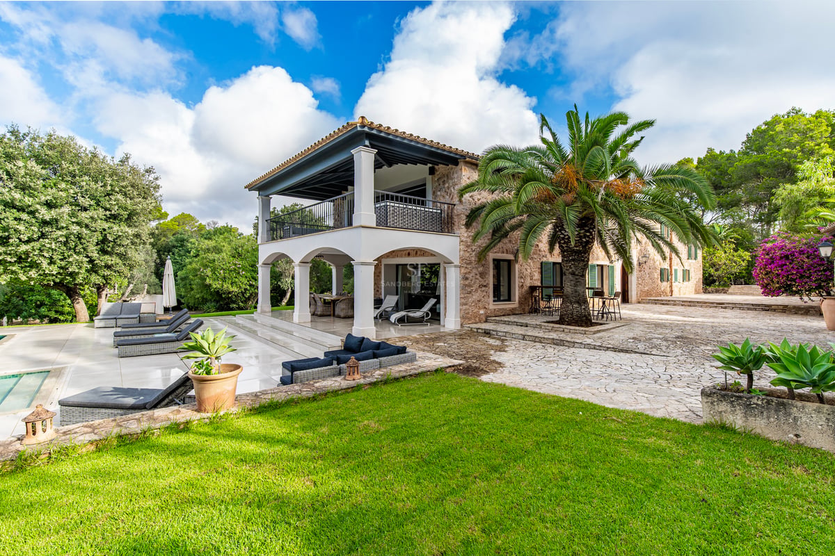 Vue extérieure d'une villa en pierre avec arches blanches, un grand palmier, des transats et un jardin paysager.