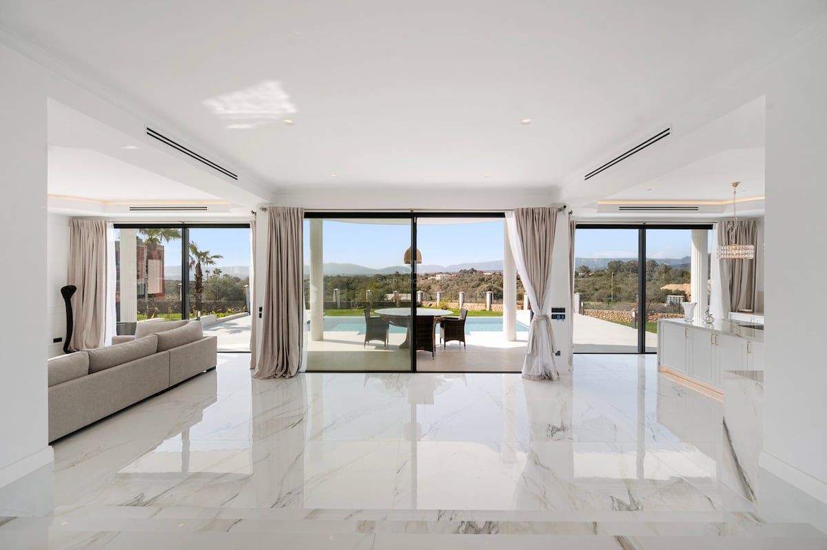 Salon lumineux avec sol en marbre blanc et baies vitrées donnant sur une piscine et les collines.
