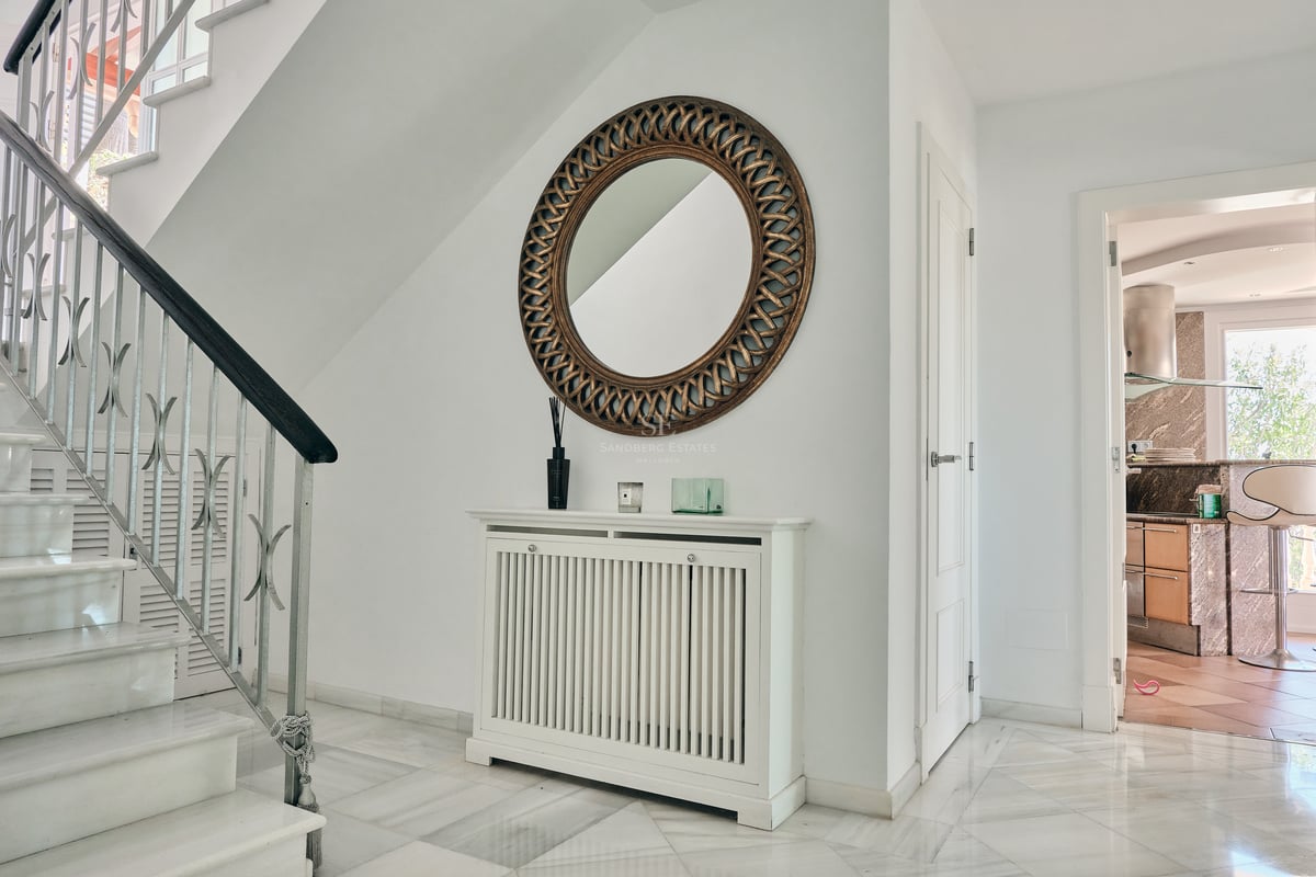 Entrée lumineuse avec escalier en marbre, cache-radiateur blanc et grand miroir circulaire orné de dorures.