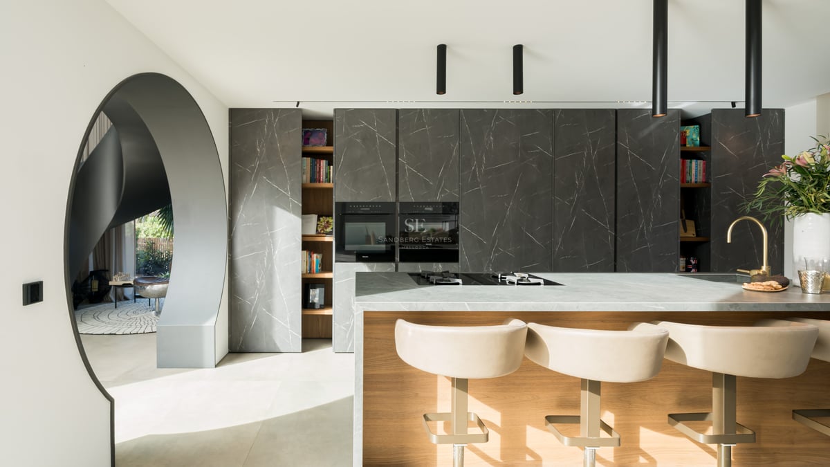 Cuisine minimaliste avec armoires en pierre sombre, îlot en bois avec tabourets blancs et appareils intégrés.