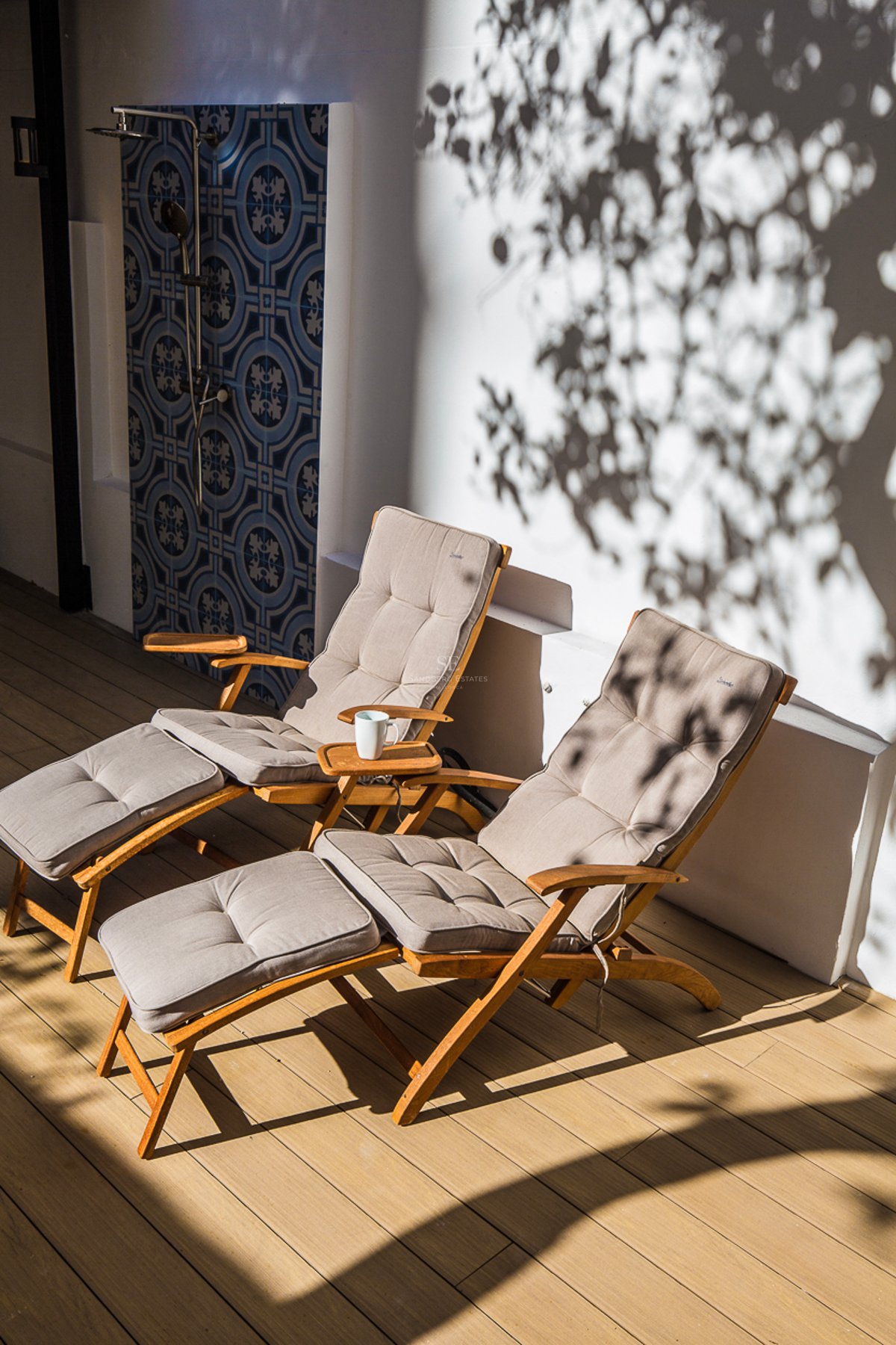Deux chaises longues en bois sur un deck à côté d'une douche extérieure carrelée de bleu avec des ombres de feuilles.