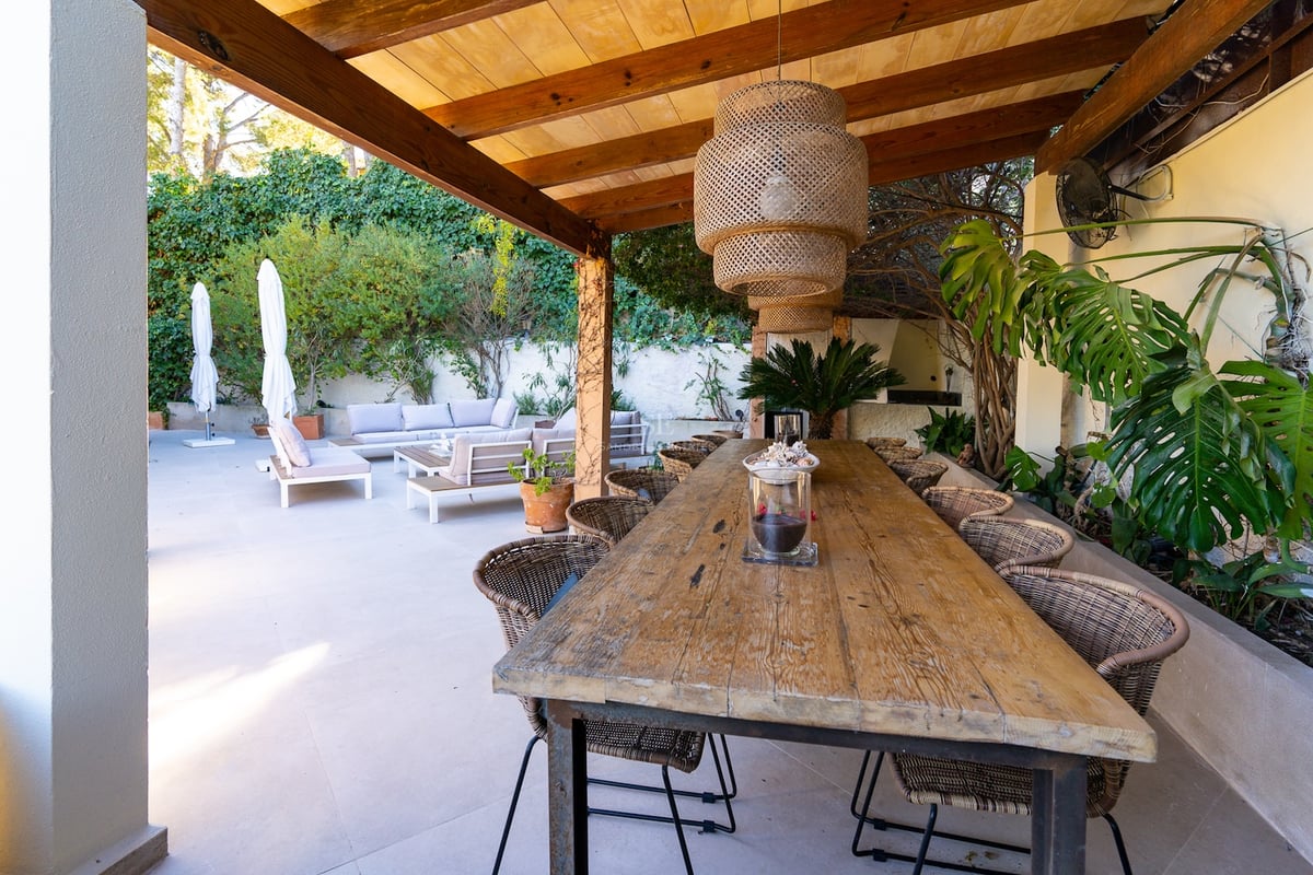 Longue table à manger en bois sous une pergola avec chaises en osier et suspension près d'un salon.