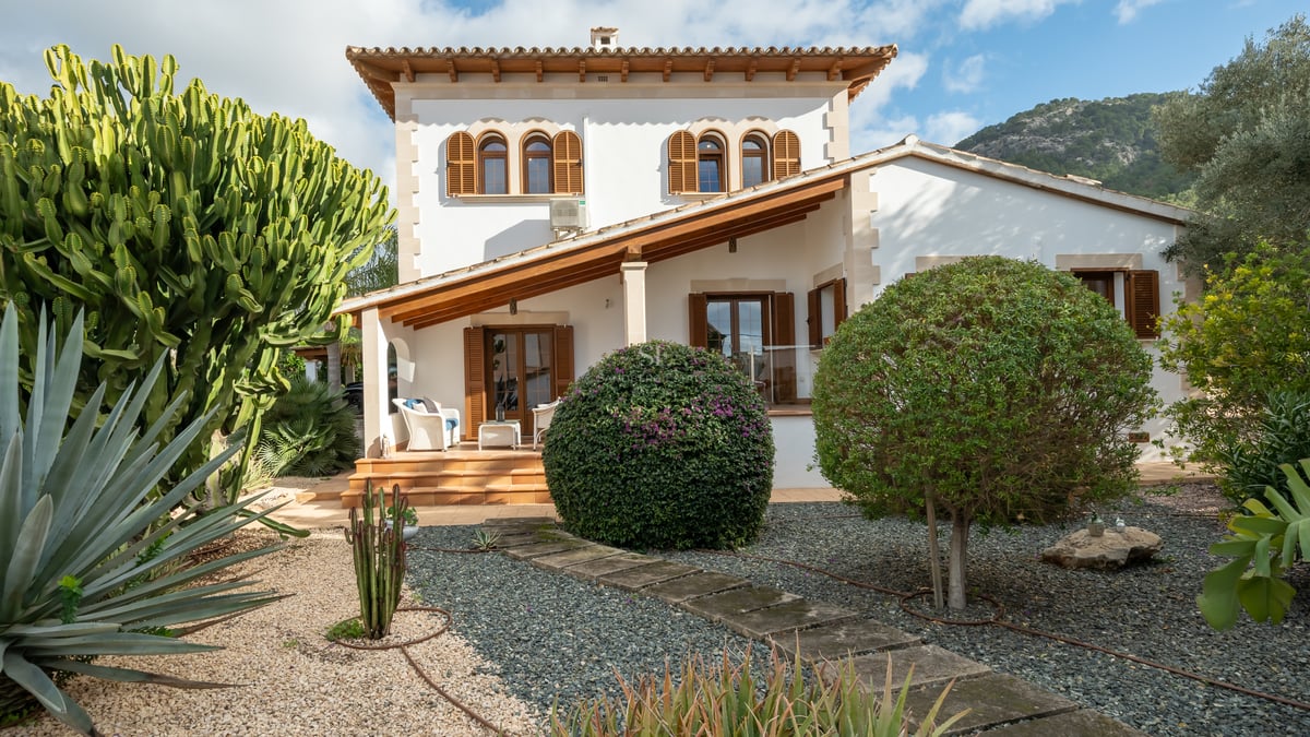 Façade de villa blanche avec volets en bois, toit en terre cuite et jardin de gravier avec cactus et agaves.