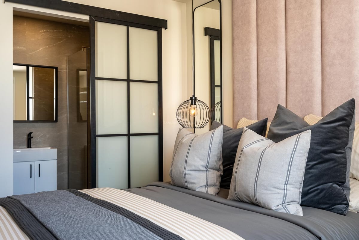 Chambre avec tête de lit en velours rose, luminaire design et porte coulissante industrielle menant à la salle de bain.
