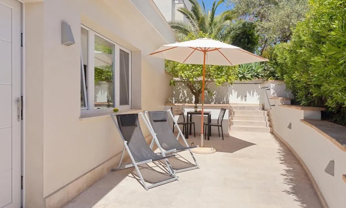 Terrasse en pierre ensoleillée avec deux chaises longues grises, un parasol blanc et un coin repas entouré de verdure.