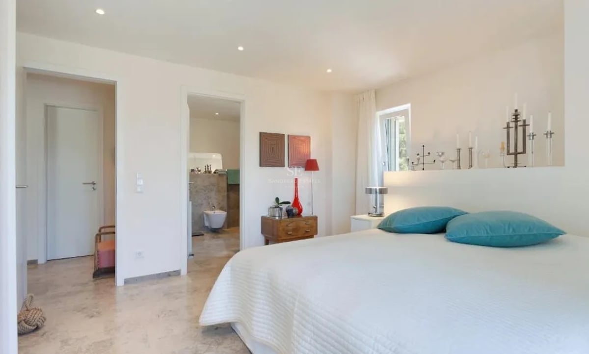 Chambre moderne avec literie blanche, coussins turquoise, sol en marbre et vue sur la salle de bains en pierre.