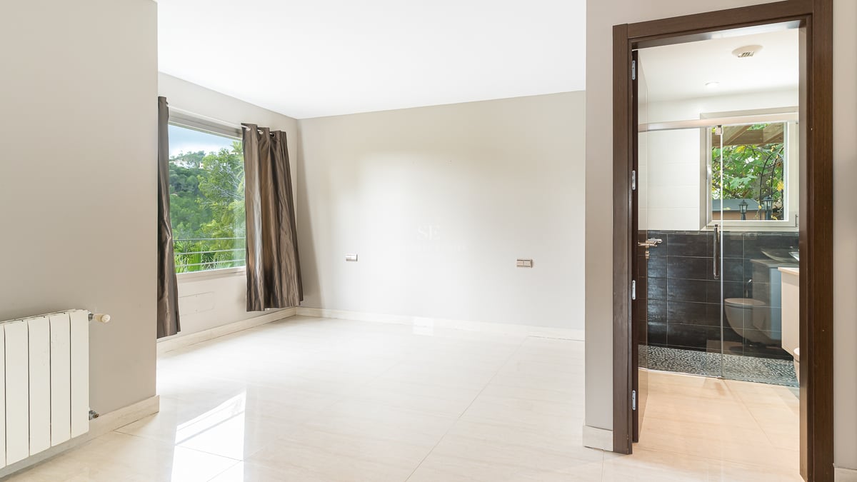 Chambre moderne vide avec sol poli, fenêtre avec rideaux et porte ouverte sur une salle de bain aux tons sombres.