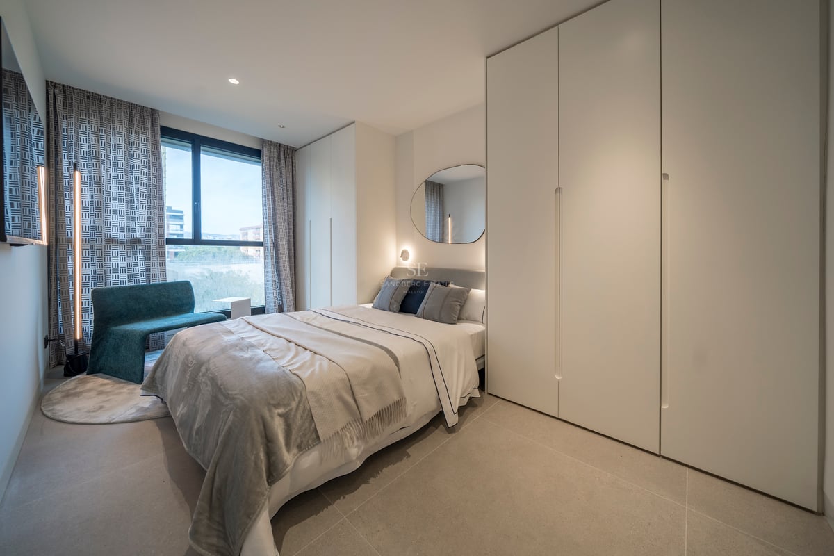 Une chambre moderne lumineuse avec un lit gris, de grandes armoires blanches, une chaise sarcelle et une grande fenêtre.