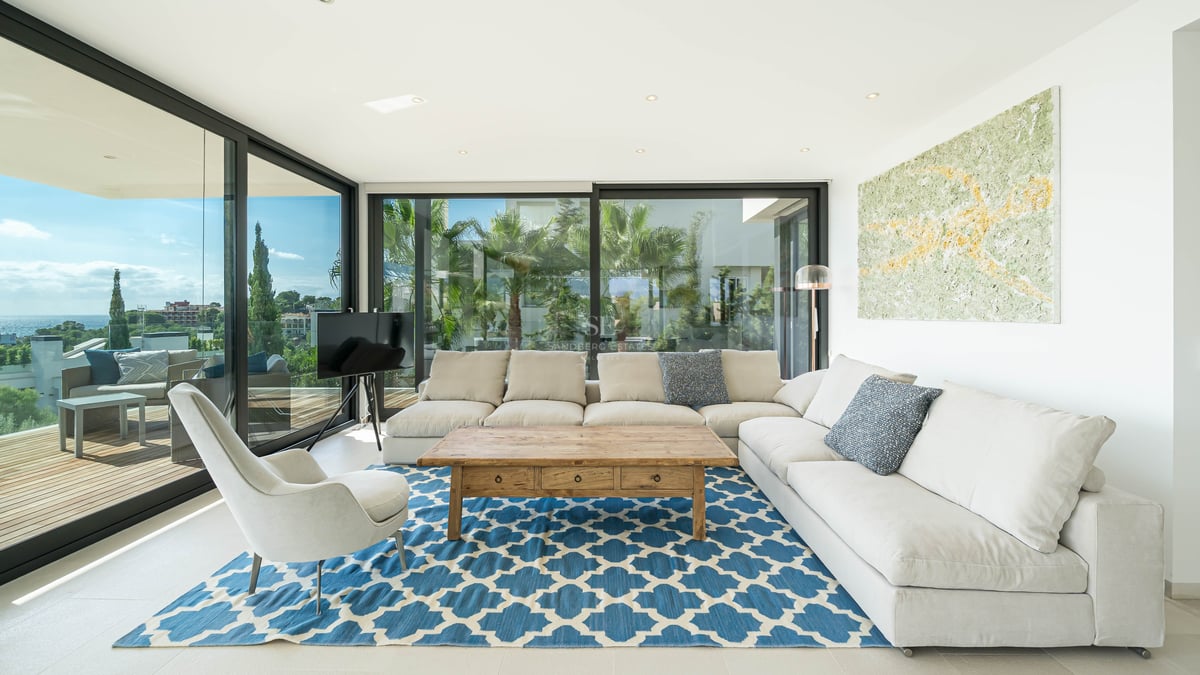 Salon moderne avec canapé en L blanc, tapis bleu à motifs et baies vitrées offrant une vue sur la côte.