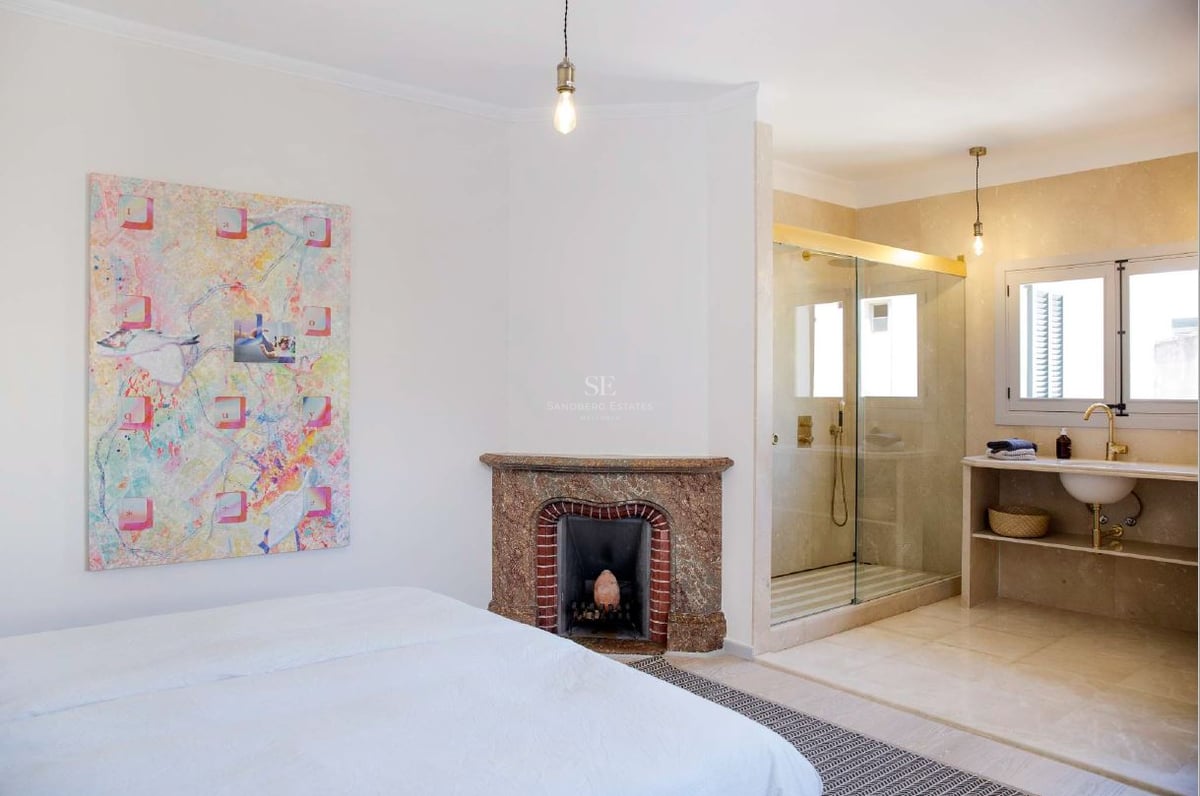 Amplio dormitorio principal con chimenea decorativa, arte moderno y baño integrado con paredes de cristal.