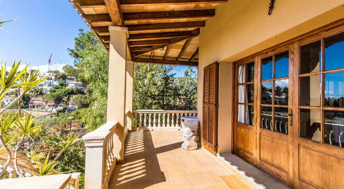 Terrasse couverte avec poutres en bois, sol carrelé et balustrade en pierre surplombant un paysage verdoyant.