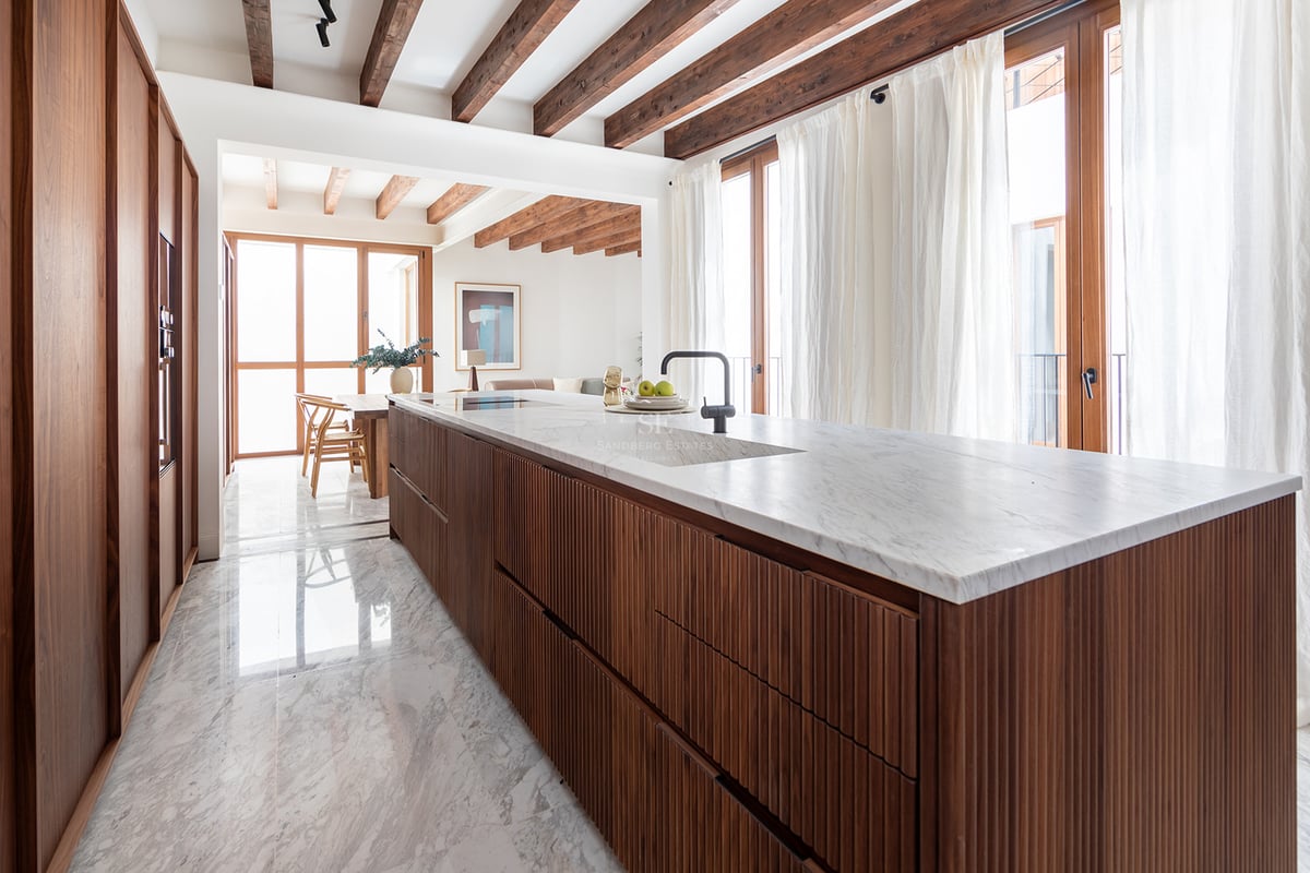 Cocina moderna con isla de madera alistonada, encimera de mármol blanco, suelos de piedra y vigas de madera en el techo.