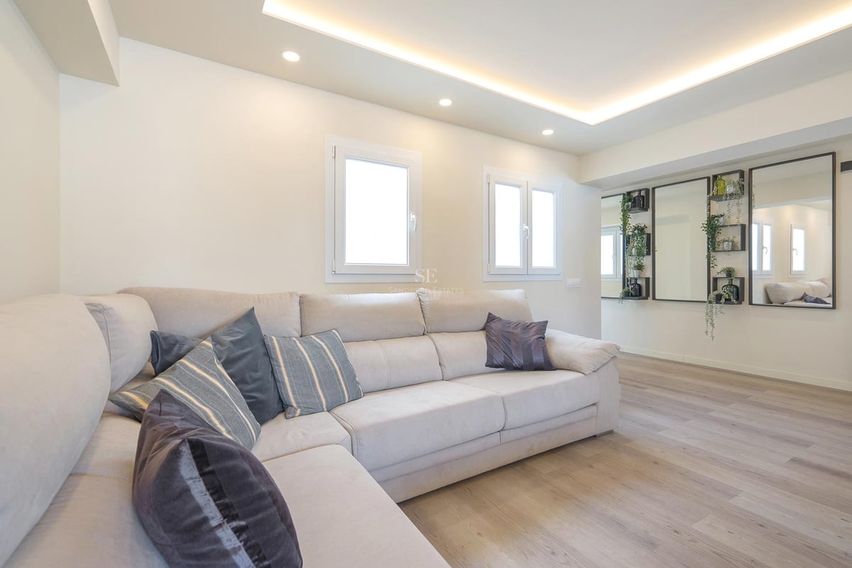 Salon moderne avec un grand canapé en L crème, parquet clair et éclairage au plafond encastré.