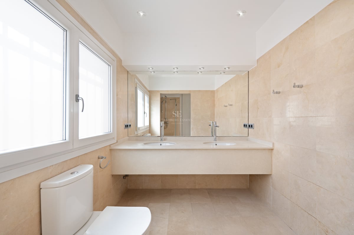 Salle de bains contemporaine avec murs en marbre crème, double vasque, grand miroir et fenêtre lumineuse.