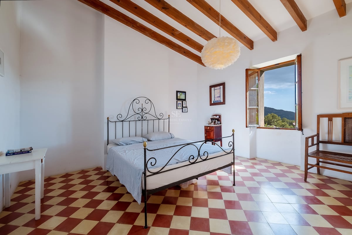 Chambre avec lit en fer forgé, sol en damier rouge et blanc, poutres en bois et fenêtre donnant sur les collines.