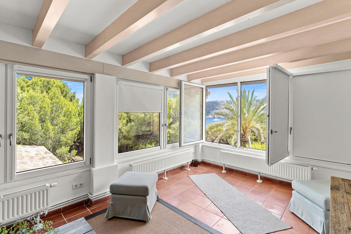 Pièce intérieure avec sol en terre cuite, poutres en bois au plafond et grandes fenêtres donnant sur des pins et la mer.