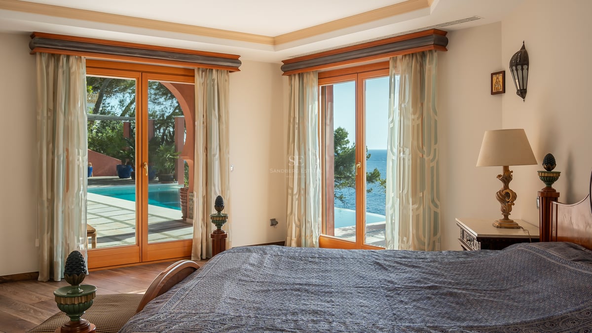 Chambre principale avec de grandes fenêtres en bois s'ouvrant sur une terrasse avec piscine et vue sur la mer.