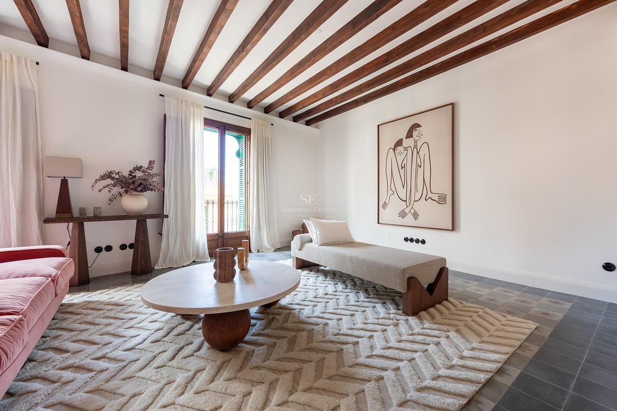 Salón luminoso con vigas de madera oscura, alfombra de espiga, mesa de travertino y acceso al balcón.