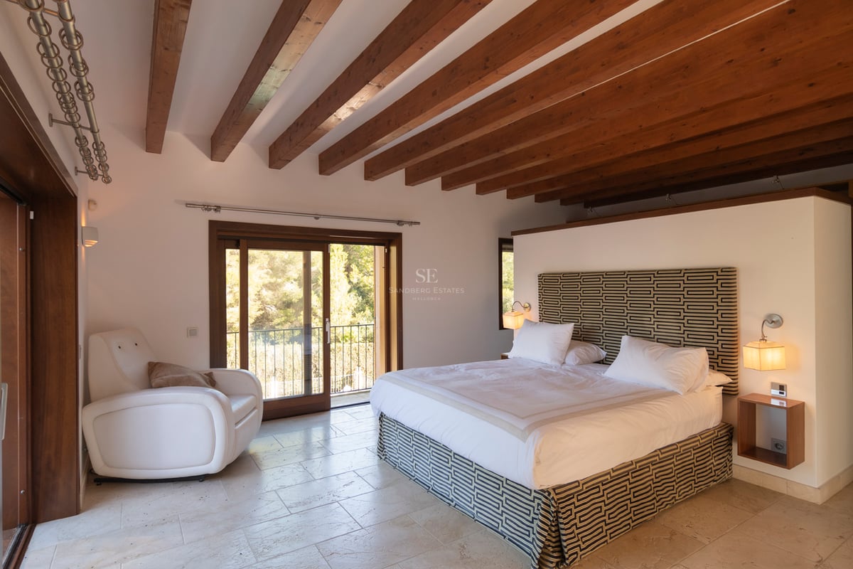 Chambre de maître avec poutres en bois au plafond, sol en pierre calcaire et accès au balcon.