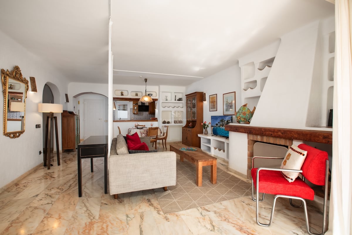 Salon lumineux avec sol en marbre poli, grande cheminée blanche, canapé gris et fauteuil rouge.