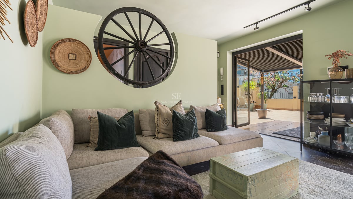 Salon chaleureux avec canapé beige, mur vert sauge et accès direct à une terrasse ensoleillée sous pergola.