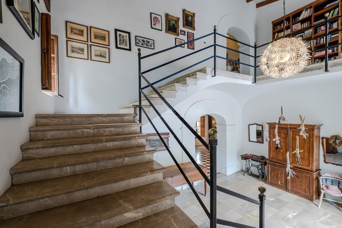 Vaste entrée avec escalier en pierre, murs blancs ornés de tableaux et bibliothèque en mezzanine.