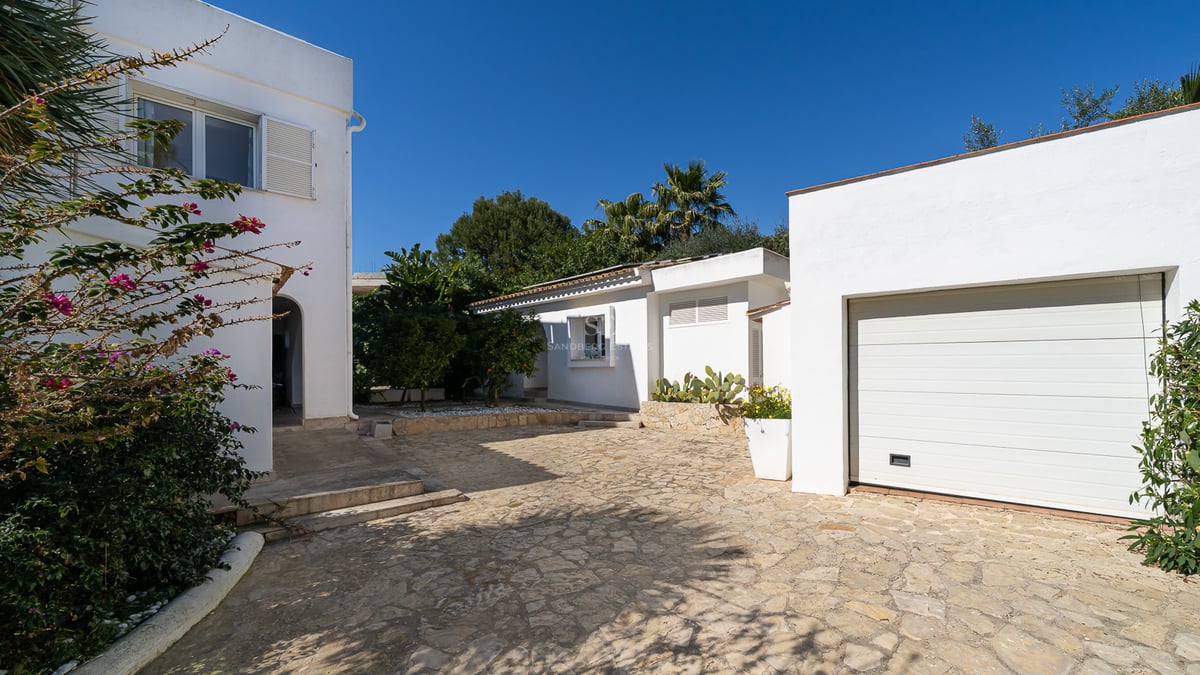 Villa méditerranéenne blanche avec une allée pavée en pierre, un garage et une végétation luxuriante sous un ciel bleu.