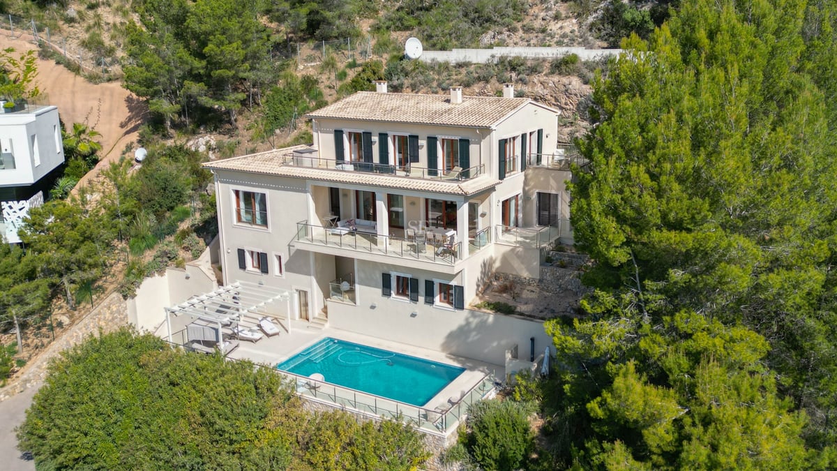 Vue aérienne d'une villa de trois étages avec piscine, terrasses en verre et toit en terre cuite entourée de pins.
