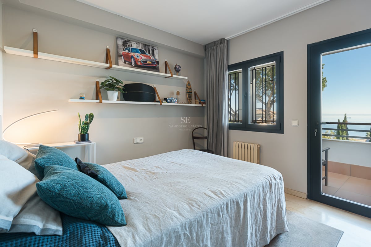 Chambre moderne aux murs gris, lit en lin, étagères murales et porte vitrée ouvrant sur un balcon avec vue mer.