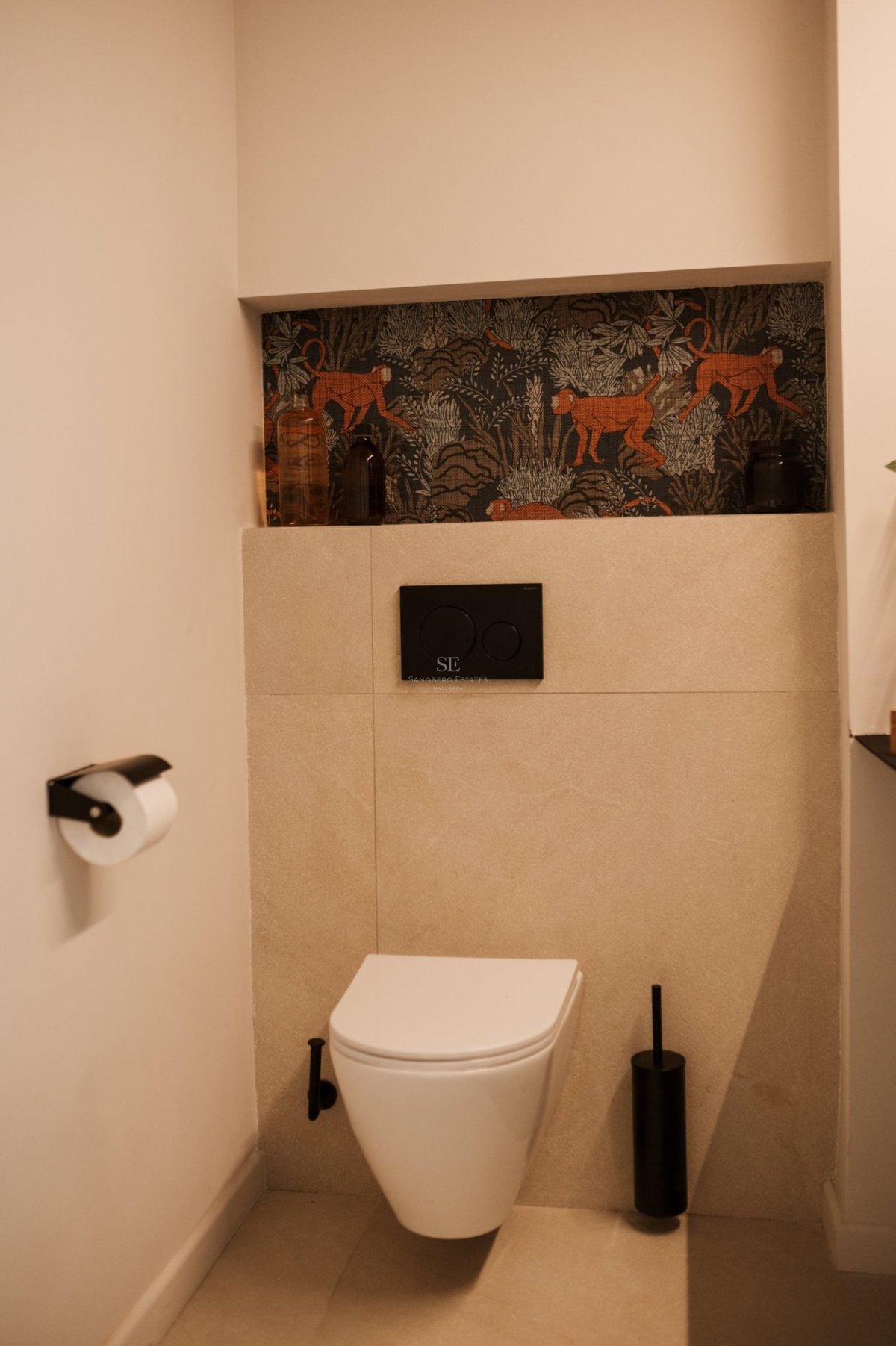 Toilettes suspendues avec plaque de déclenchement noire, carrelage beige et niche décorative à motif de singes.