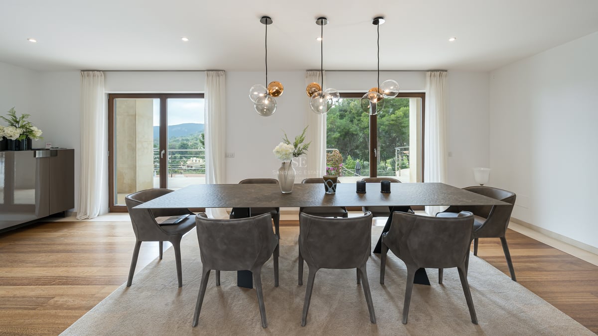 Salle à manger moderne avec table texturée, chaises grises et suspensions en verre devant de grandes baies vitrées.