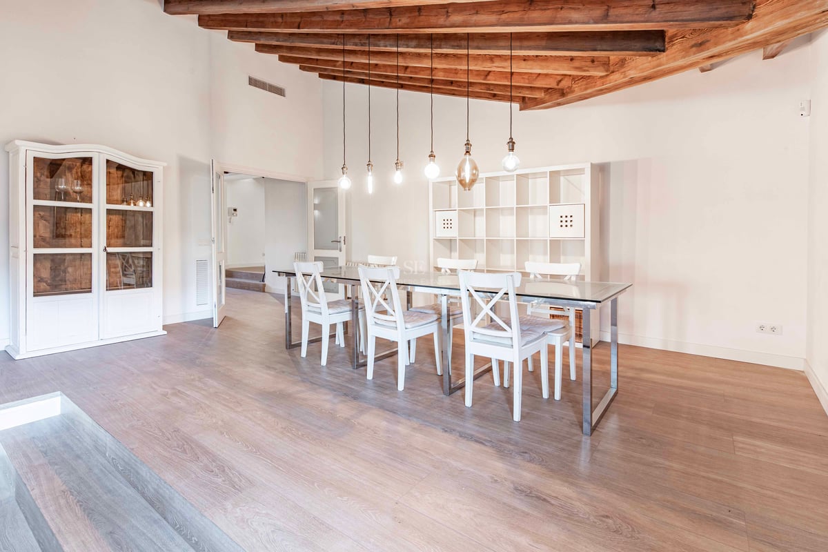 Une salle à manger spacieuse avec table en verre, chaises blanches et hauts plafonds avec poutres rustiques.