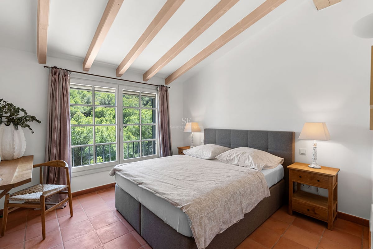 Chambre avec lit double gris, poutres apparentes, sol en terre cuite et vue sur la forêt.