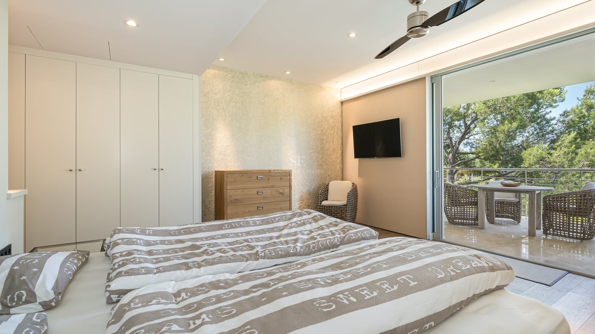 Chambre moderne avec placards blancs, commode en bois et baies vitrées ouvrant sur une terrasse ombragée par des pins.