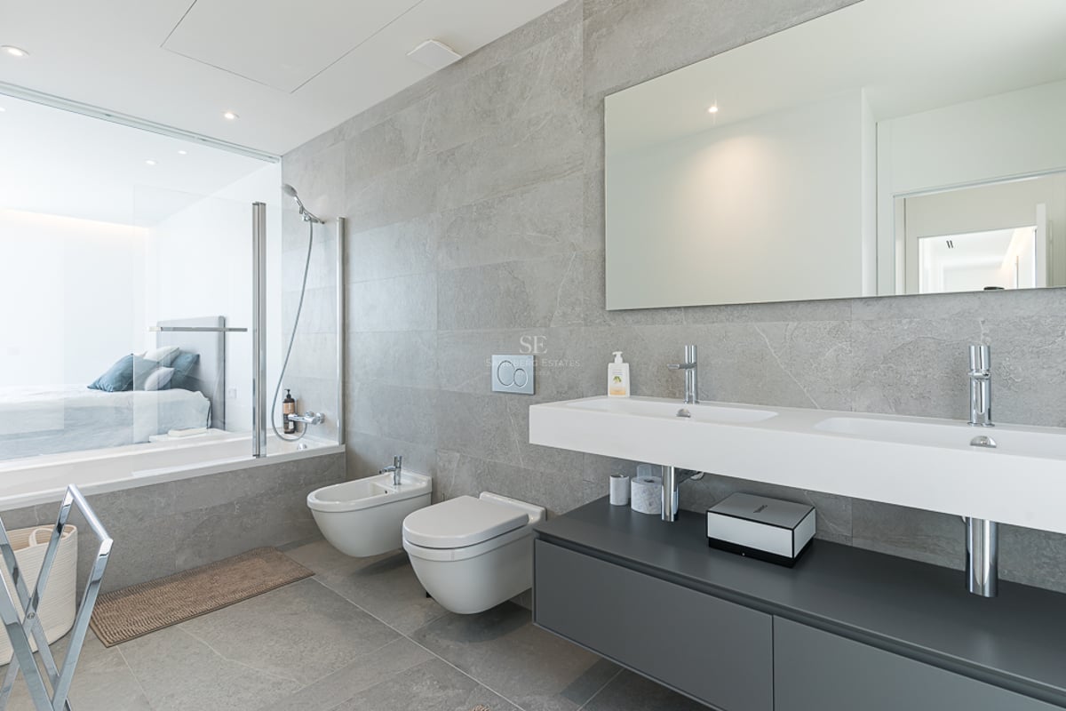 Salle de bain moderne avec carrelage gris, double vasque, WC suspendus et paroi vitrée donnant sur la chambre.