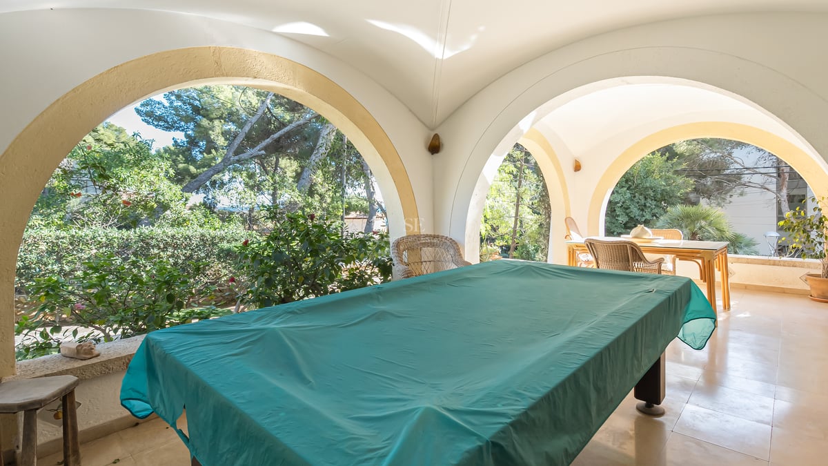 Grande terrasse couverte avec arches blanches, table de billard sous bâche verte et vue sur le jardin.