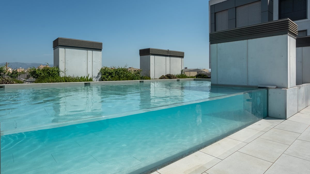 Piscine moderne sur le toit avec paroi en verre transparent et carrelage en pierre grise sous un ciel bleu.