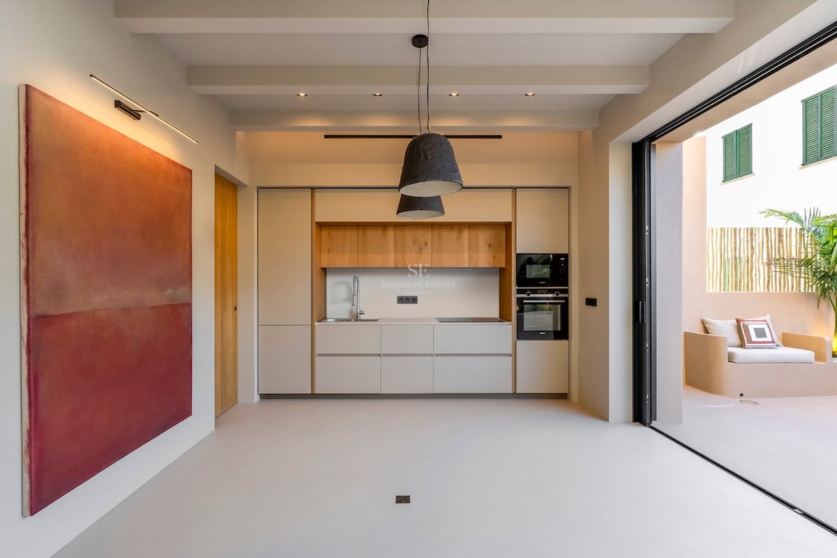 Cuisine minimaliste avec accents en bois clair, grand tableau rouge et baies vitrées ouvrant sur un patio ensoleillé.