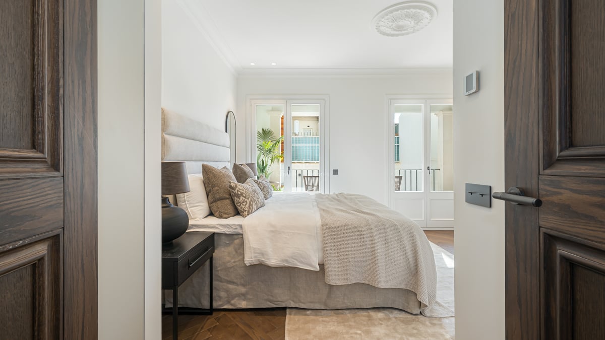 Bright bedroom seen through dark wood doors, featuring a large bed, herringbone floors, and French balcony doors.
